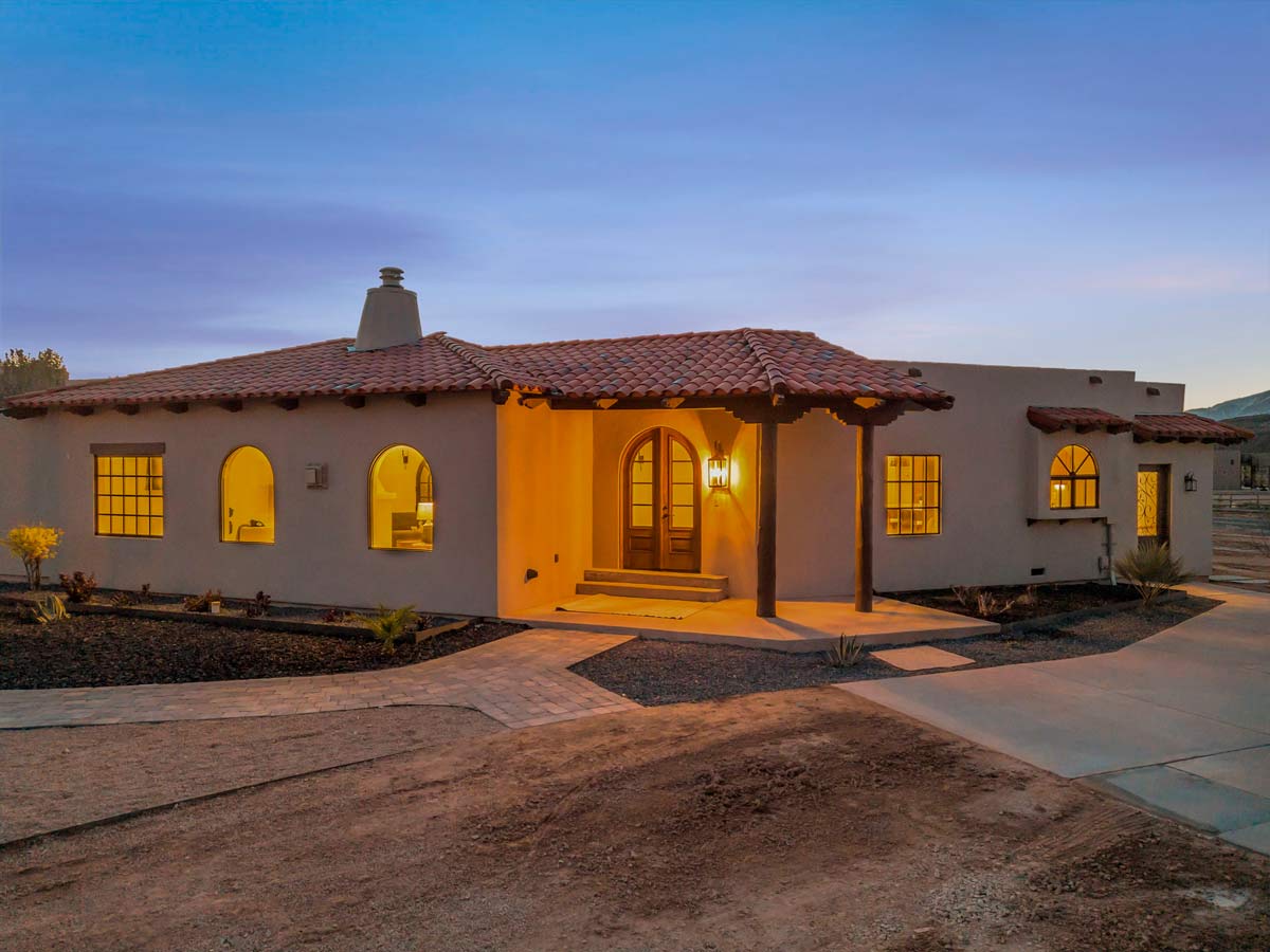 A Spanish-style house at dusk with adobe walls and a red-tiled roof. Warm light emanates from the windows and front porch, highlighting the curved arches and wooden door. Surrounding landscaping is visible, with a paved walkway leading to the entrance.