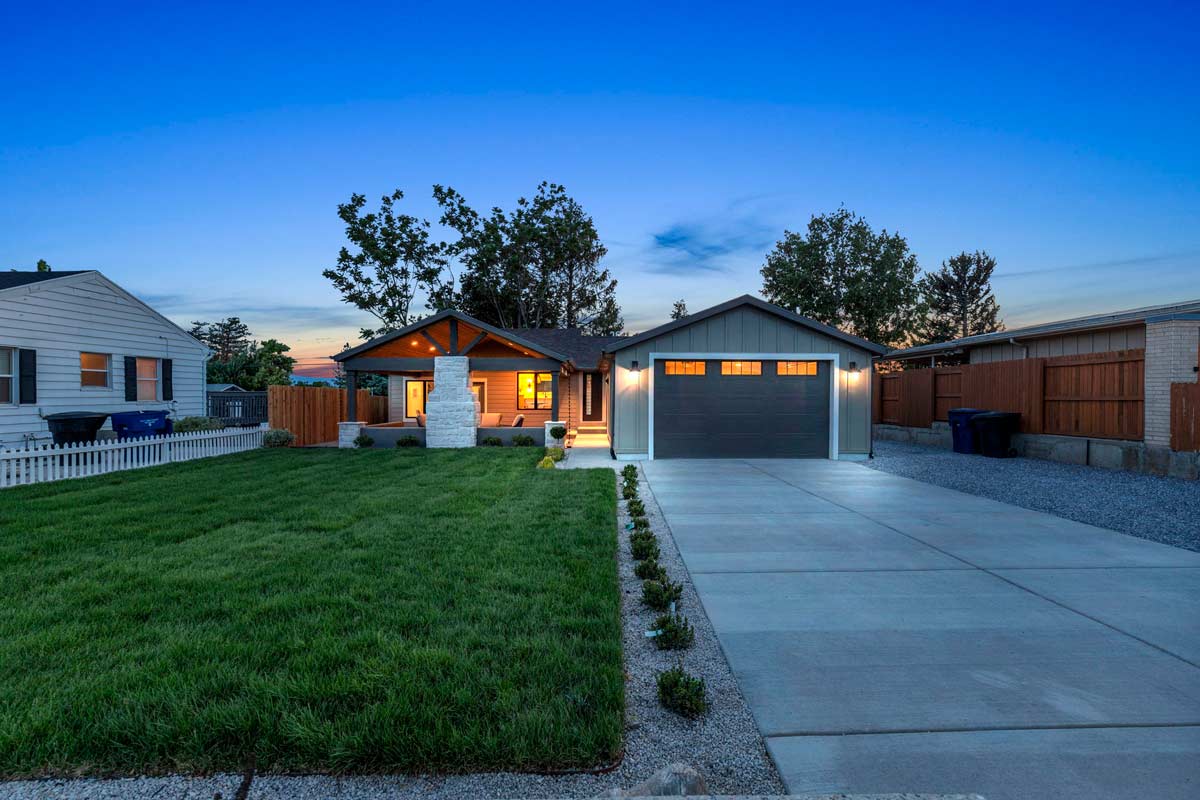 A modern single-story house with a well-manicured lawn and spacious driveway at dusk. The homes windows and lights emit a warm glow. The driveway is bordered by small plants, and the sky is clear, transitioning to night.