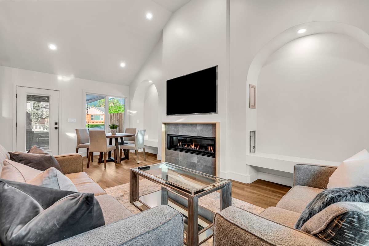 A modern living room featuring a fireplace beneath a wall-mounted TV, surrounded by two gray armchairs and a sofa. A glass coffee table sits in the center. In the back, a dining table with four chairs and a window showcasing greenery. Bright lighting.