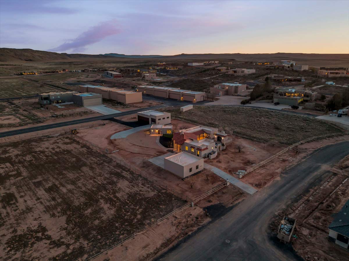 Aerial view of a residential area at dusk. The foreground features a house with a red roof and surrounding buildings. The landscape is desert-like with sparse vegetation, and the sky is transitioning from sunset to evening.