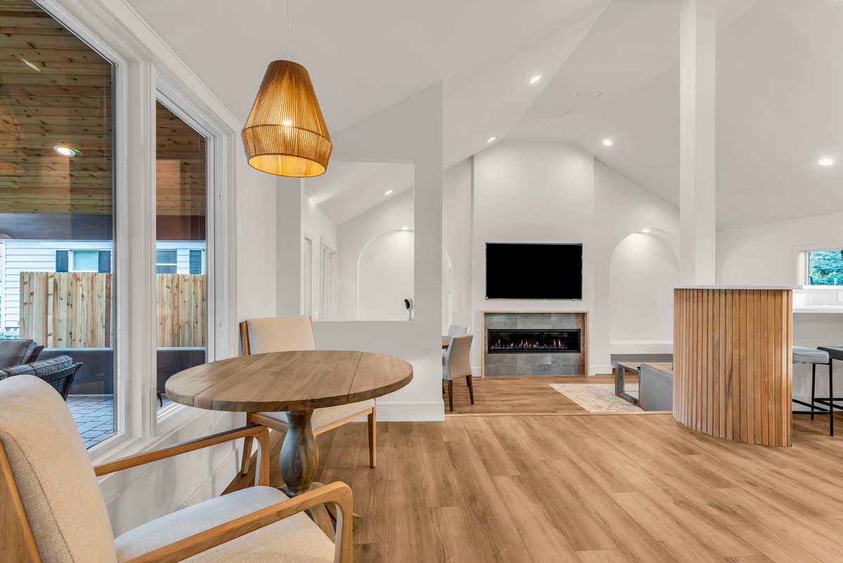 Modern living room with a high ceiling, featuring a wooden floor, a round dining table with chairs, and a pendant light. A fireplace with a TV above it is on the far wall, alongside a curved wooden partition and large windows overlooking a deck.