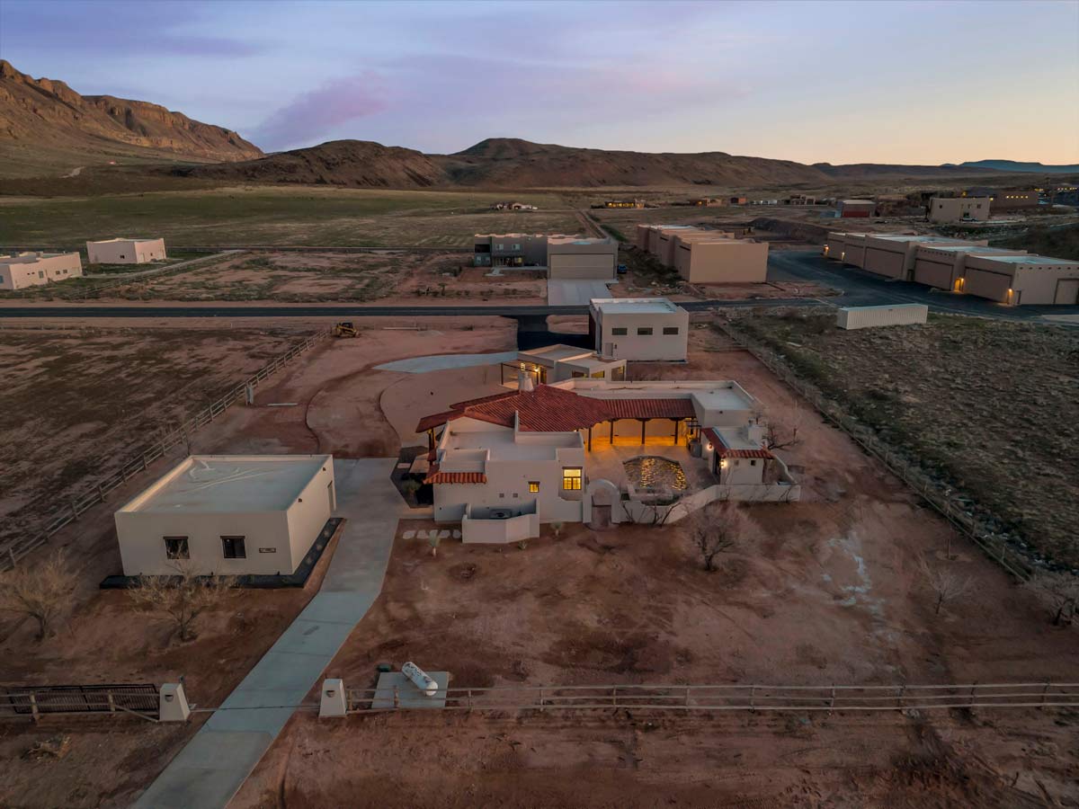 Aerial view of a desert property at sunset. The main house has a circular design with a red-tiled roof, surrounded by smaller buildings, a driveway, and a fence. The hilly landscape extends in the background under a colorful sky.