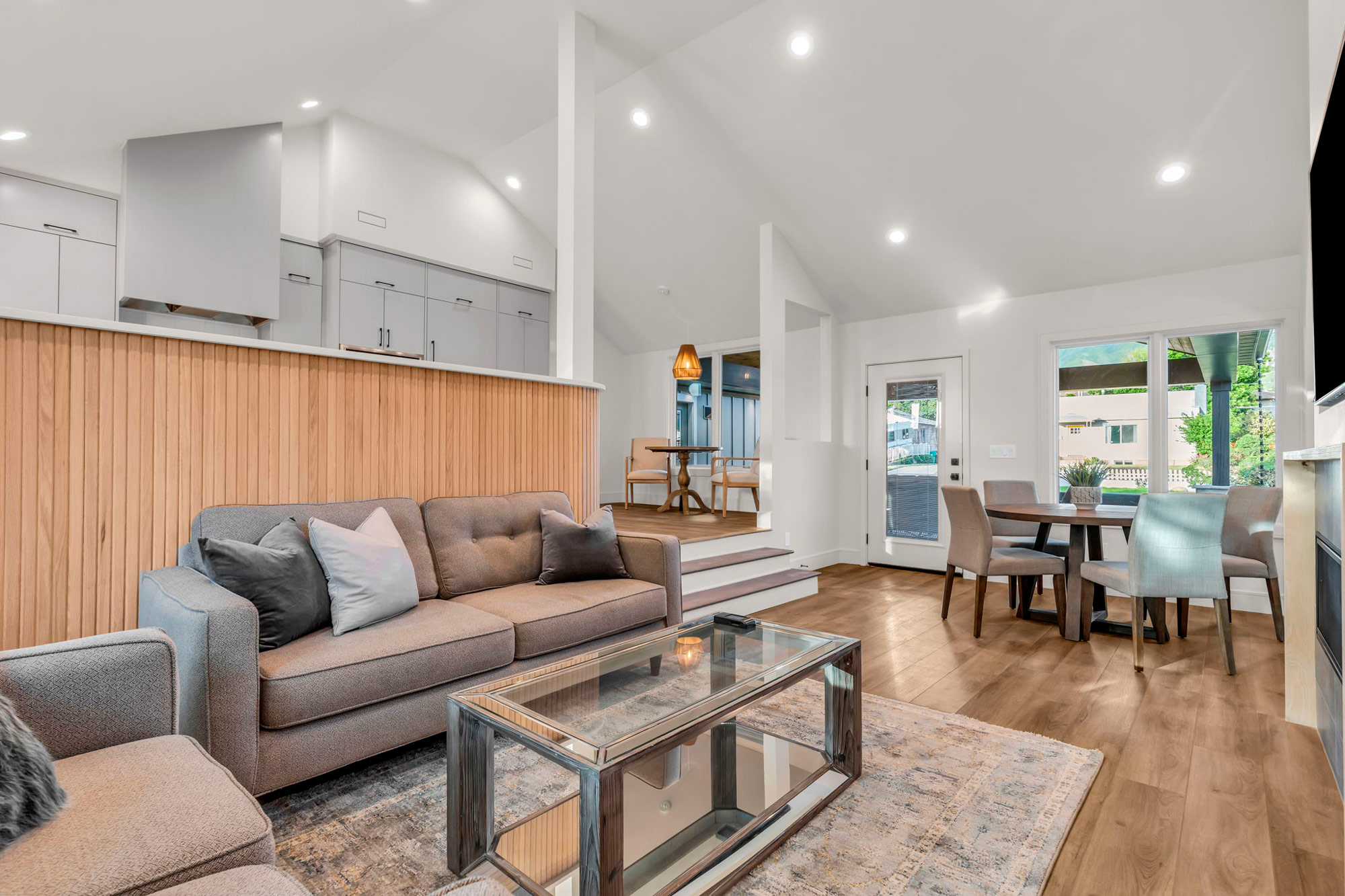 Modern open-concept living room and kitchen with hardwood floors. The space features a gray sofa, glass coffee table, wooden accent wall, and dining table by a large window. Bright lighting and a blend of contemporary decor create a welcoming atmosphere.