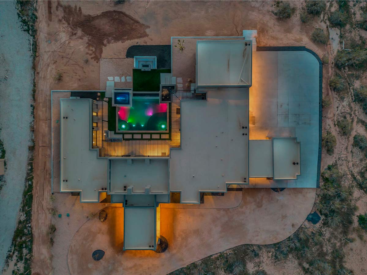 Aerial view of a modern, flat-roofed building surrounded by desert terrain. The rooftop features colorful lights and a small pool area. Warm lights illuminate the buildings exterior, creating a contrast with the natural surroundings.