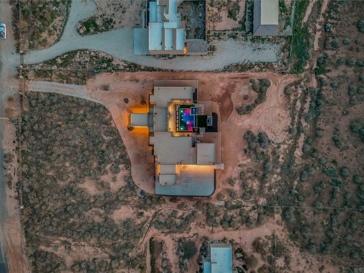 Aerial view of a modern building in a desert landscape. The structure has a boxy design with a central area brightly illuminated. Surrounding the building are dirt paths and patches of sparse vegetation.