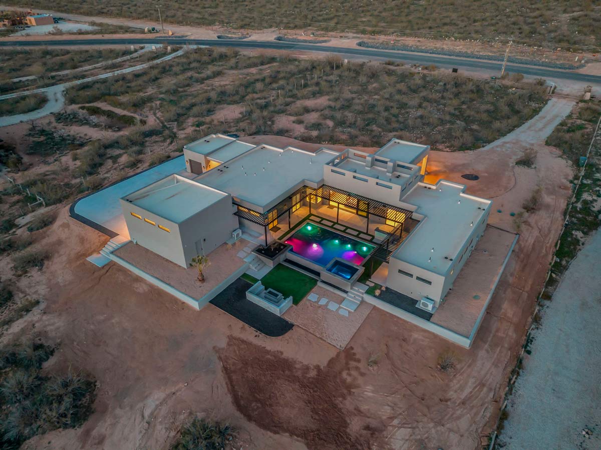 Aerial view of a modern, luxurious house in a desert landscape. The house features a large pool with colorful lights, a patio area, and minimalist architecture. Surrounding sandy terrain and sparse vegetation are visible.