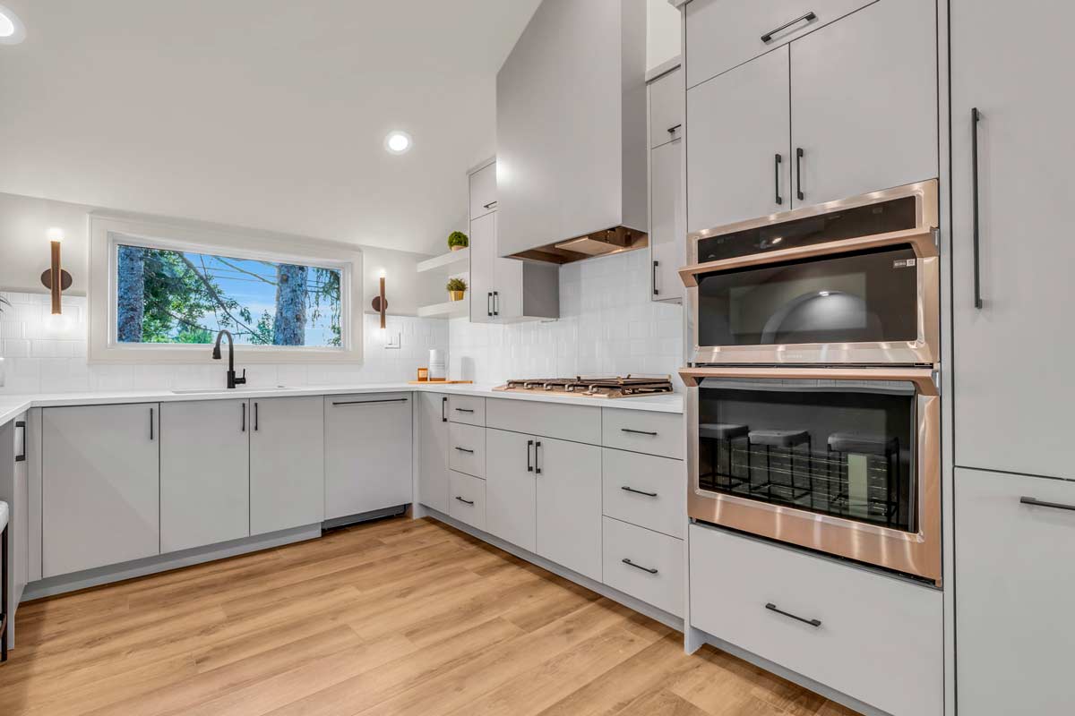 A modern kitchen featuring light gray cabinetry, a double oven, and a gas cooktop. The room has wooden flooring and a large window allowing natural light. Minimalist decor with built-in dishwasher and sleek wall lights complement the clean design.