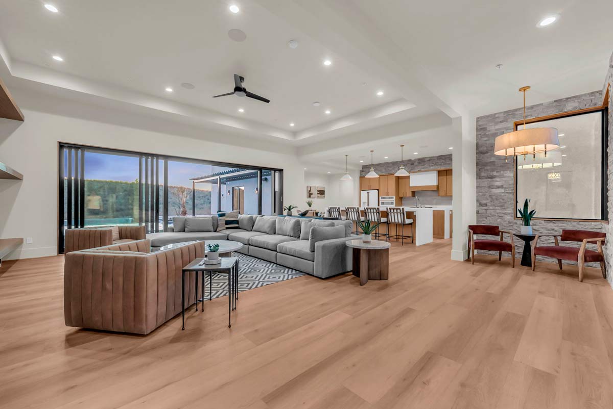 Spacious modern living room with wooden floors, featuring a large sectional sofa, two armchairs, and a patterned rug. Open layout includes a kitchen with bar stools. Large sliding doors reveal an outdoor area. Ceiling fan and recessed lighting are present.