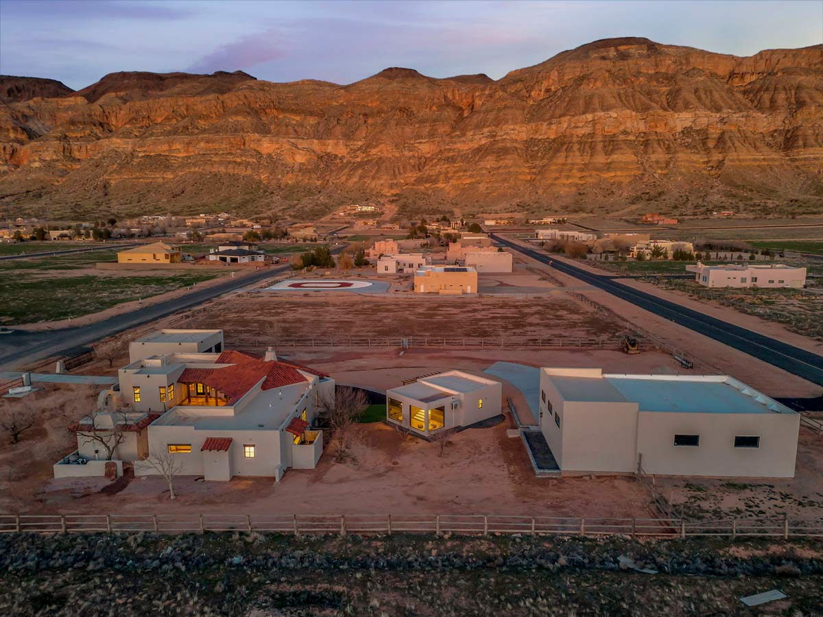 Aerial view of a desert residential area at sunset, featuring modern-style houses with flat roofs. The landscape includes rugged mountains in the background, and the setting sun casts warm tones over the scene. A helicopter pad is visible.