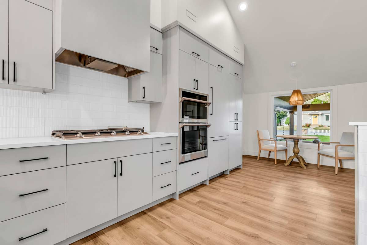 Modern kitchen with light gray cabinets, stainless steel oven and range, and wood flooring. A small dining area with a round table and two chairs is by a window. Bright and spacious.
