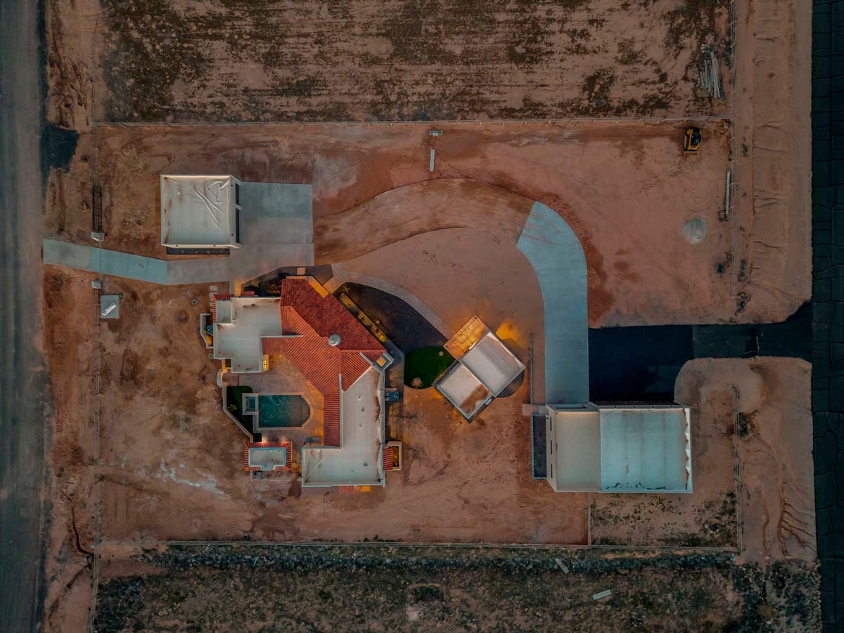 Aerial view of a house under construction in a desert area. The home features a red-tiled roof, a curved driveway, and nearby structures. Surrounding the property are barren, dirt-filled plots and a partially paved road.