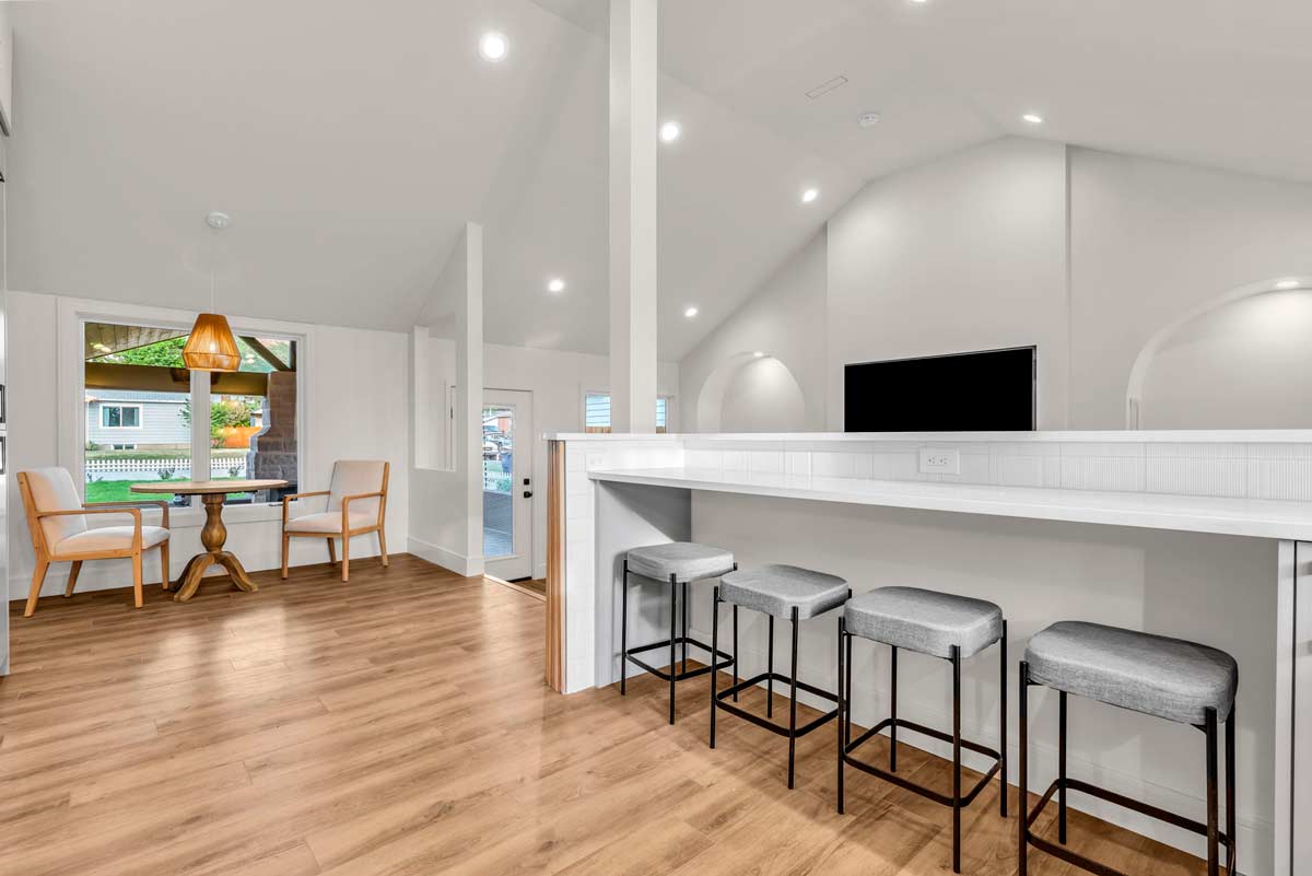 Modern open-concept living space with wooden flooring, high ceilings, and white walls. Features a bar counter with gray stools, a TV on the wall, and a small dining area with a round table and chairs under a hanging light. Large windows allow natural light in.
