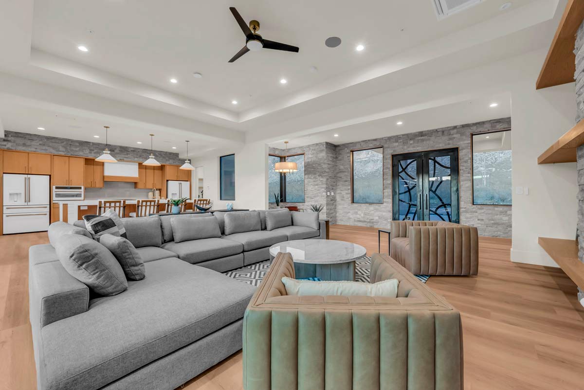 Modern open-plan living room with a large gray sectional sofa and two beige armchairs. A round coffee table sits on a patterned rug. The kitchen in the background features wooden cabinetry and a breakfast bar with white stools.