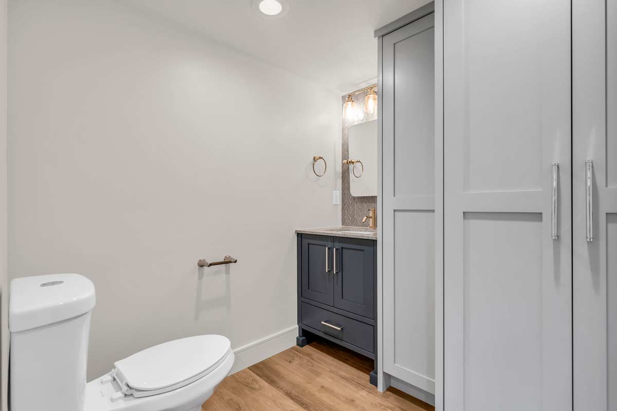 A modern bathroom featuring a white toilet, a wooden floor, a blue vanity with gold fixtures, and a rectangular mirror. Two round towel holders are mounted on the wall beside the mirror, with a cabinet on the right side.
