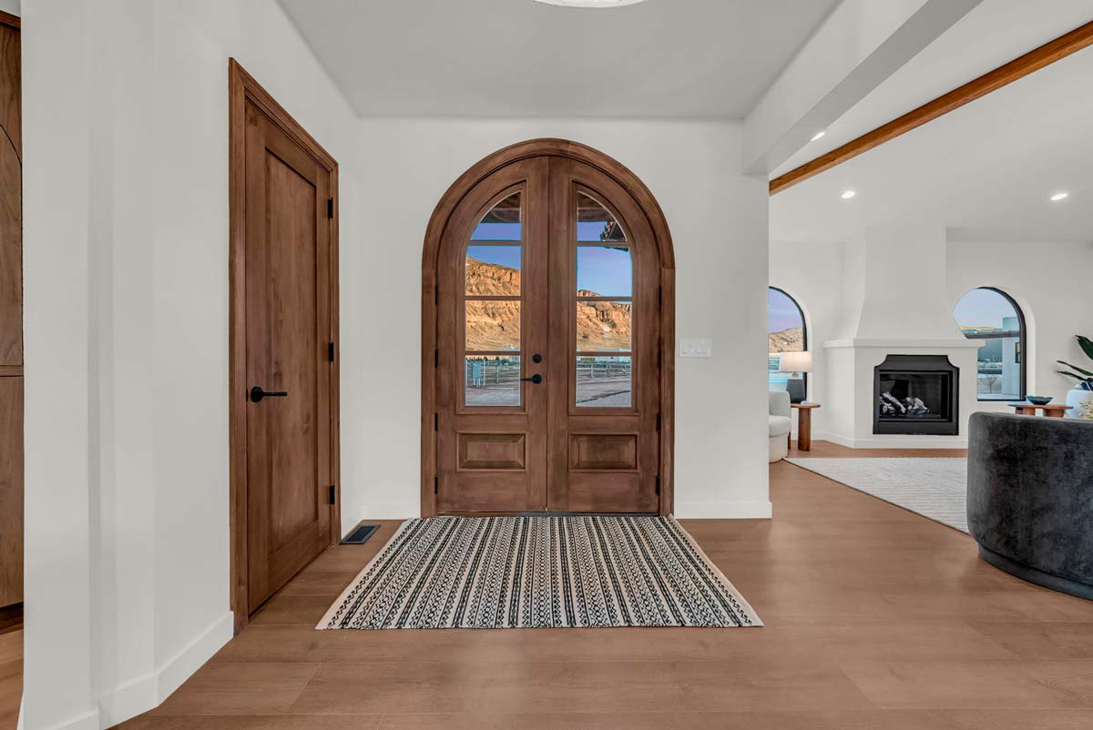 A wooden arched front door with glass panels is at the center, flanked by white walls. A patterned rug lies on the floor. Through the door, theres a view of mountains and water. To the right, a living area features a fireplace and large windows.