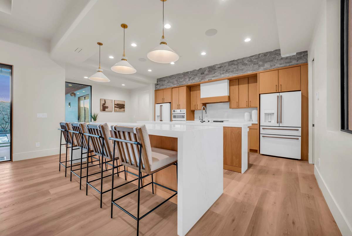 Modern kitchen with a large white island, bar stools, and pendant lighting. Wooden cabinets and stainless steel appliances line the back wall. Hardwood floors and a window overlooking a scenic view complete the space.