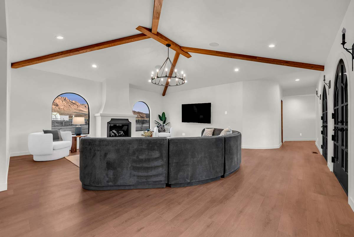 Spacious living room with white walls, a curved gray sofa, a black fireplace, and a wall-mounted TV. Features a wooden beam ceiling with a circular chandelier. Arched windows provide natural light and outdoor views. Light wood flooring throughout.