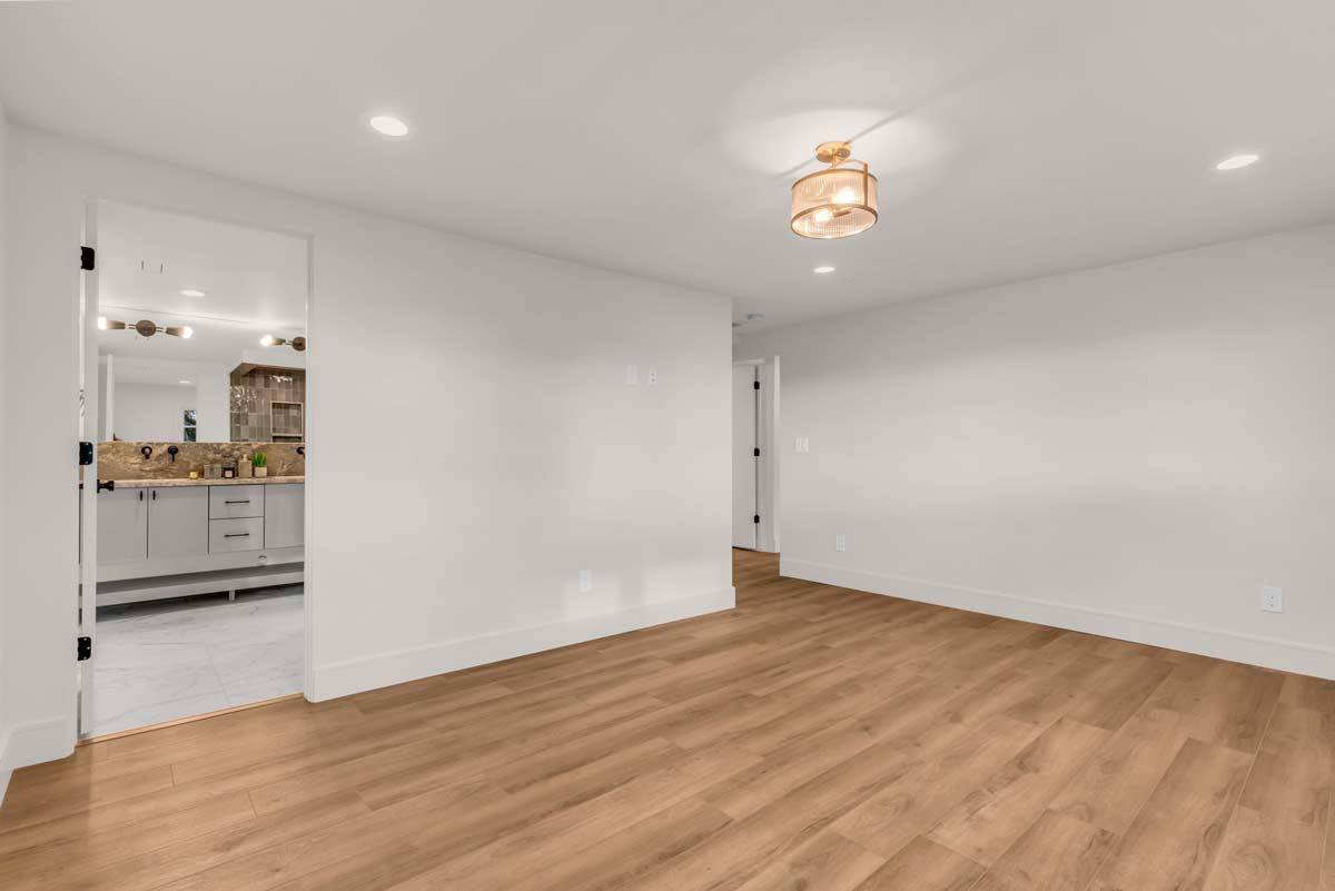 An empty room with light wood flooring and white walls. A ceiling light fixture hangs overhead. The open door reveals a bathroom with a double sink vanity and a wall mirror. Recessed lighting is visible on the ceiling.