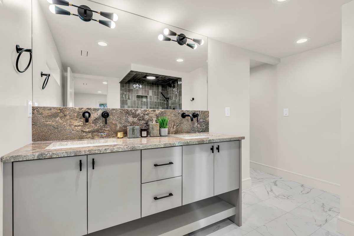 Modern bathroom with a wide mirror above a two-sink vanity featuring a granite countertop. The space is lit by sleek black fixtures. The floor is tiled in a light marble pattern, and a shower area is visible in the background.