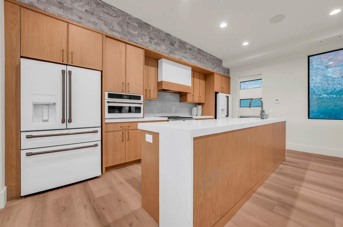 Modern kitchen with wooden cabinets and a large white island. Features include a double-door refrigerator, built-in oven, microwave, and sink. Grey backsplash tiles and wood flooring provide a contemporary look, with a large window offering natural light.