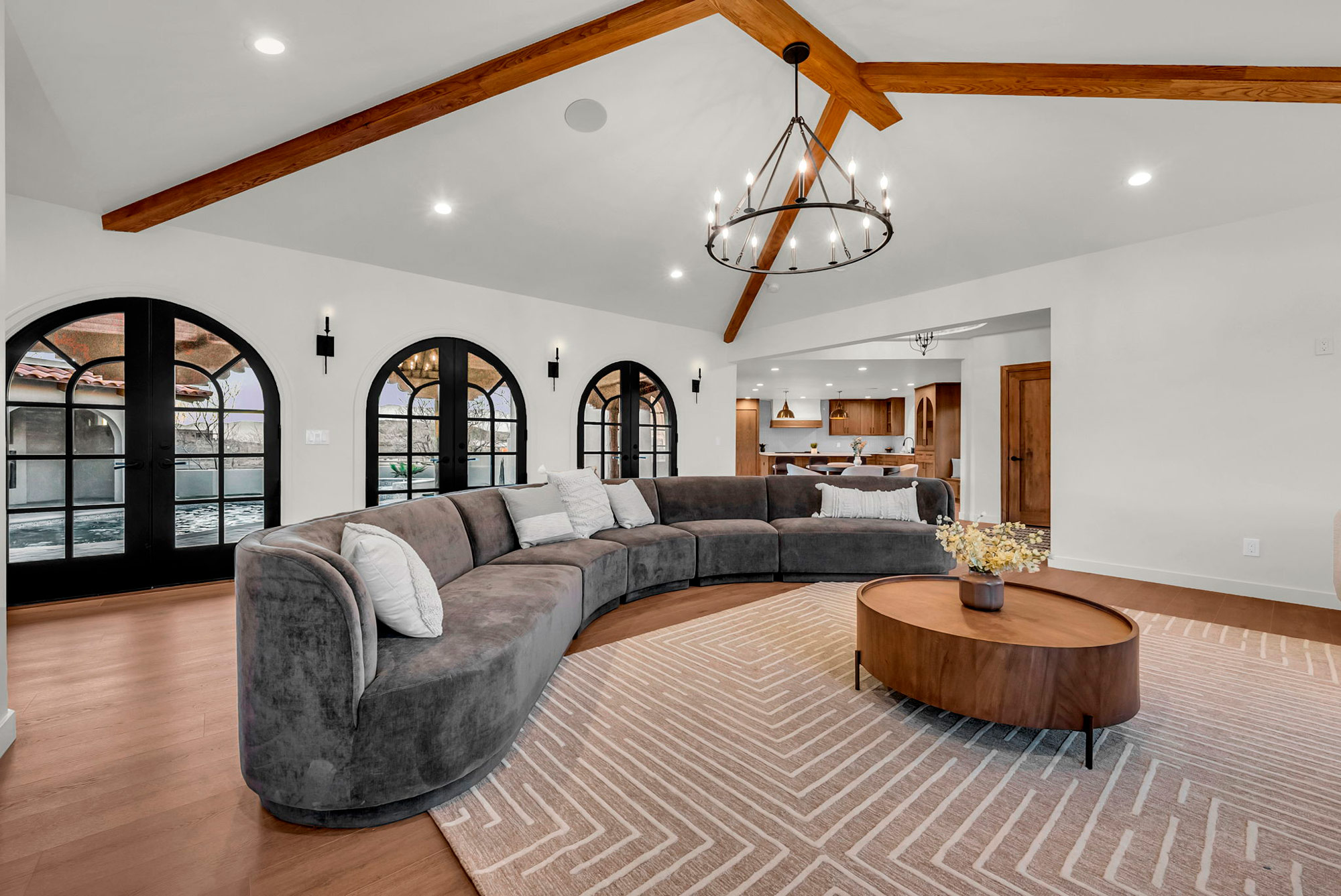 Spacious living room with a curved gray sofa, wooden coffee table, and geometric rug. Exposed ceiling beams and a modern chandelier add elegance. Three arched doors lead outside, and a kitchen is visible in the background.