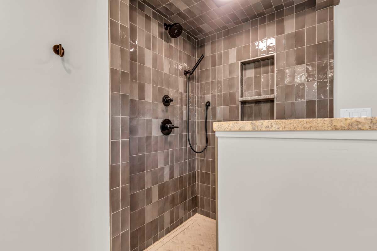 Shower area with dark gray tiles and two showerheads, including a handheld option. A built-in shelf is present on the wall. Part of a countertop with an electrical switch is visible in the foreground.