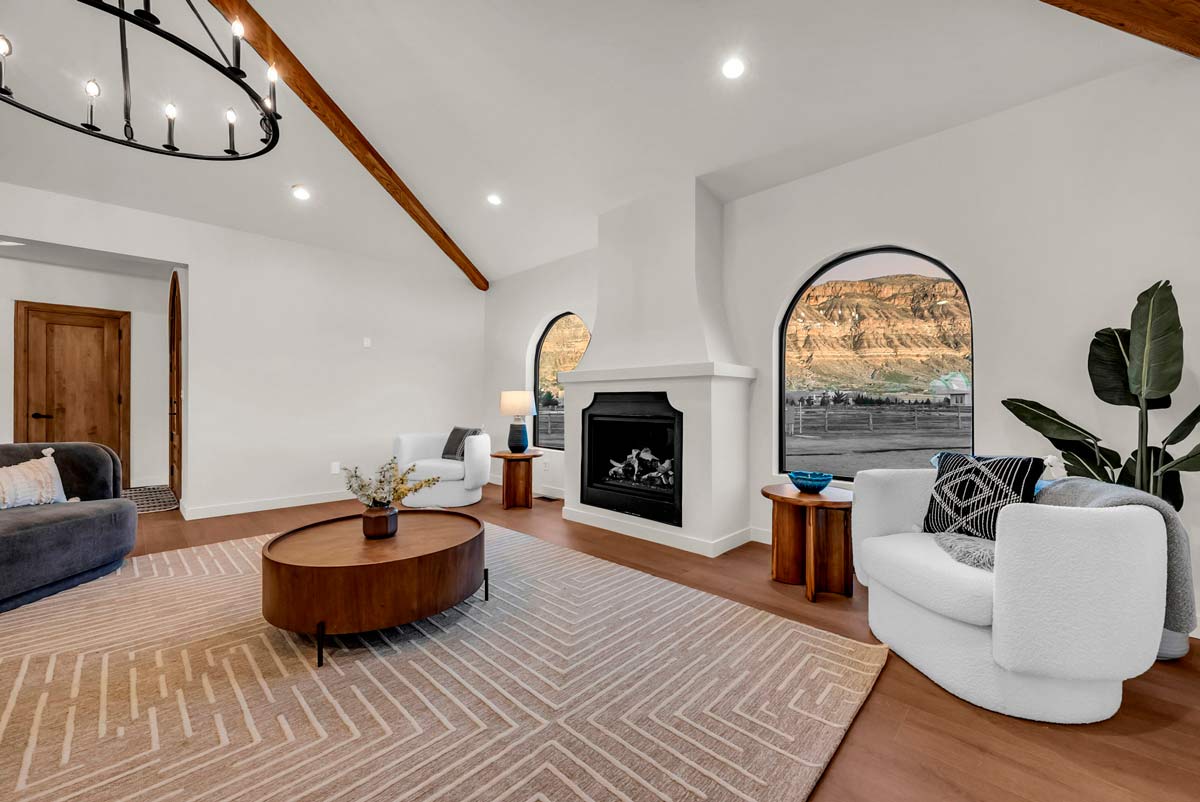 Spacious living room with a white and wood theme. Features a circular coffee table, modern fireplace, cozy seating, large patterned rug, and arched windows showcasing a scenic landscape. Indoor plant and stylish decor complete the inviting space.