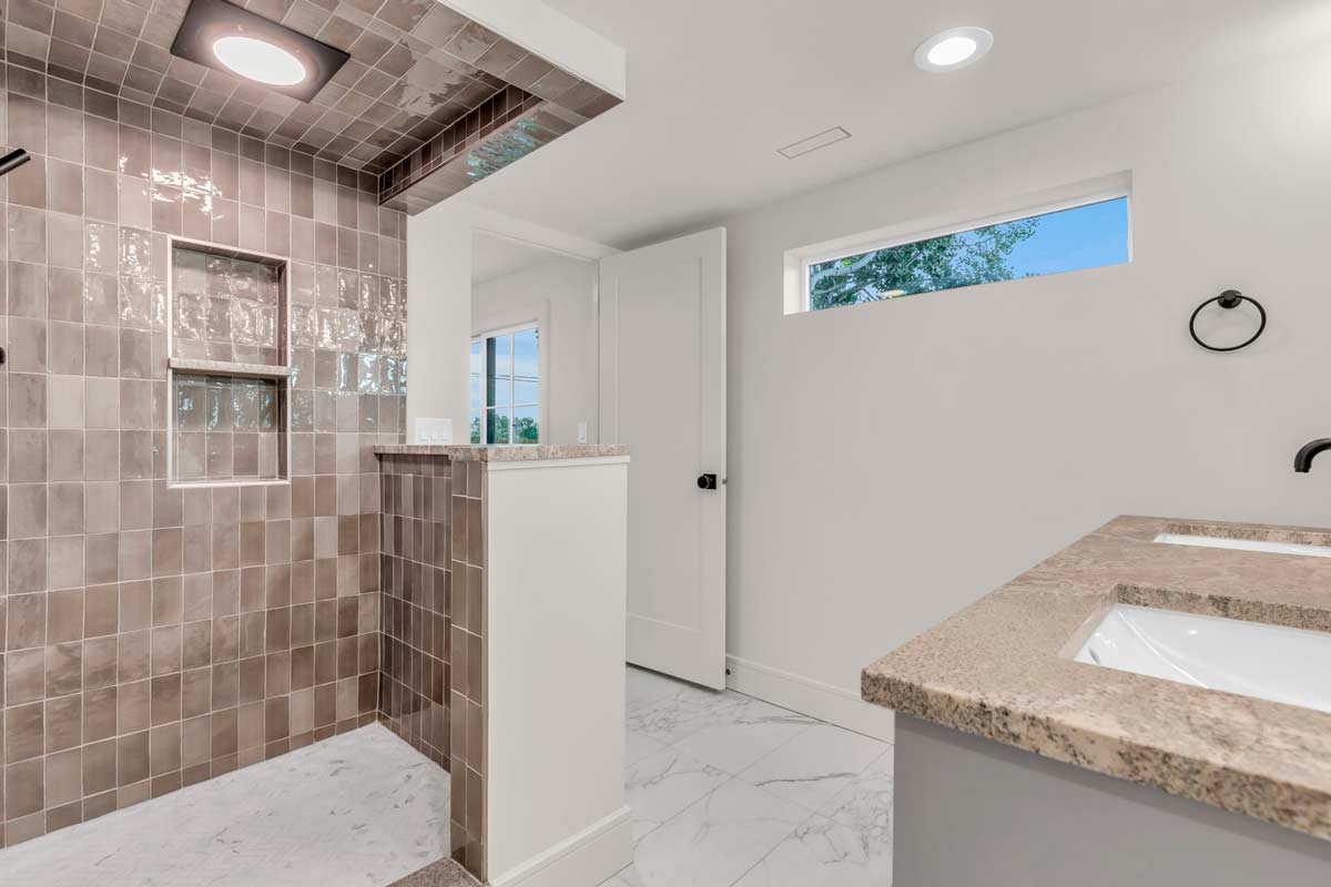 Modern bathroom featuring a walk-in shower with brown tiles, small wall niches, a high showerhead, and a half-wall partition. Marble floor, beige countertop, rectangular sink, and a large mirror. A narrow window provides natural light.