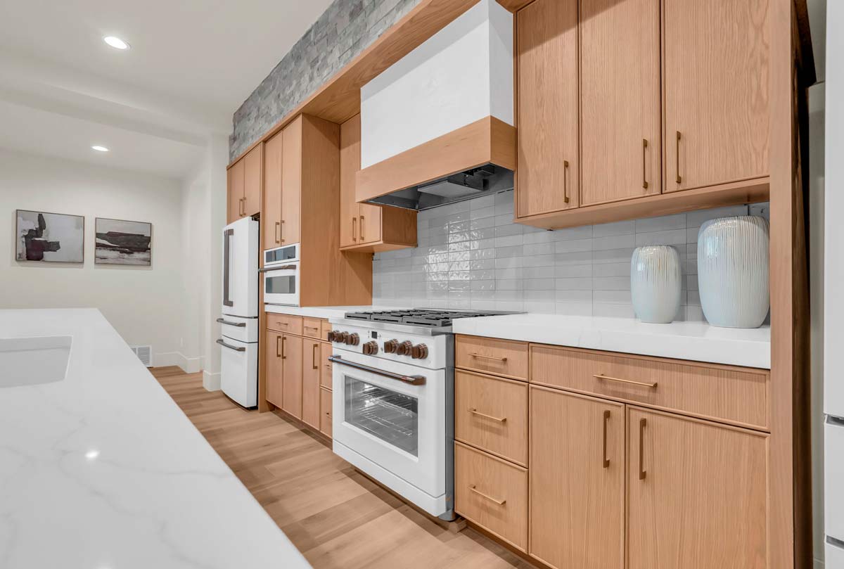 A modern kitchen with light wood cabinets and white countertops. Theres a white stove and oven, a tiled backsplash, a range hood, and two large decorative vases. Recessed lighting and two wall artworks enhance the space.