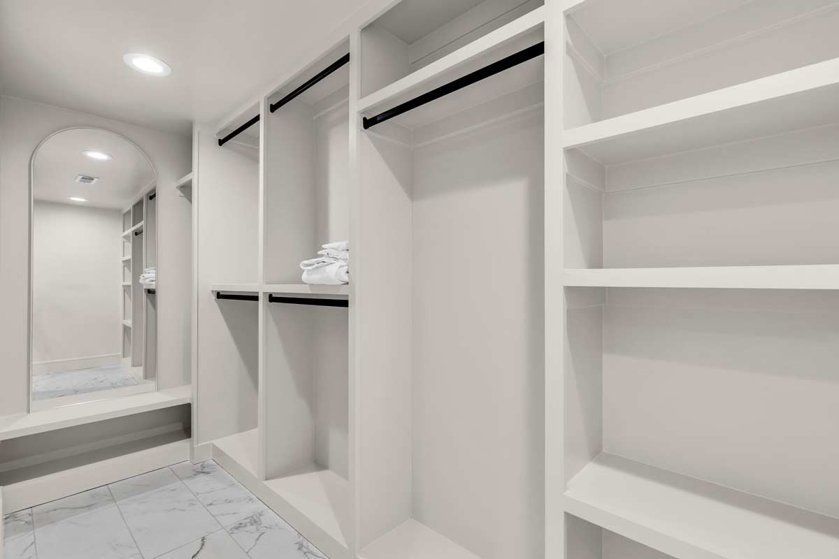 A spacious, empty walk-in closet with white shelving and hanging rods. Neatly folded white towels are on one shelf. The closet features white walls, a tiled floor, and a full-length mirror with an arched top on the back wall.