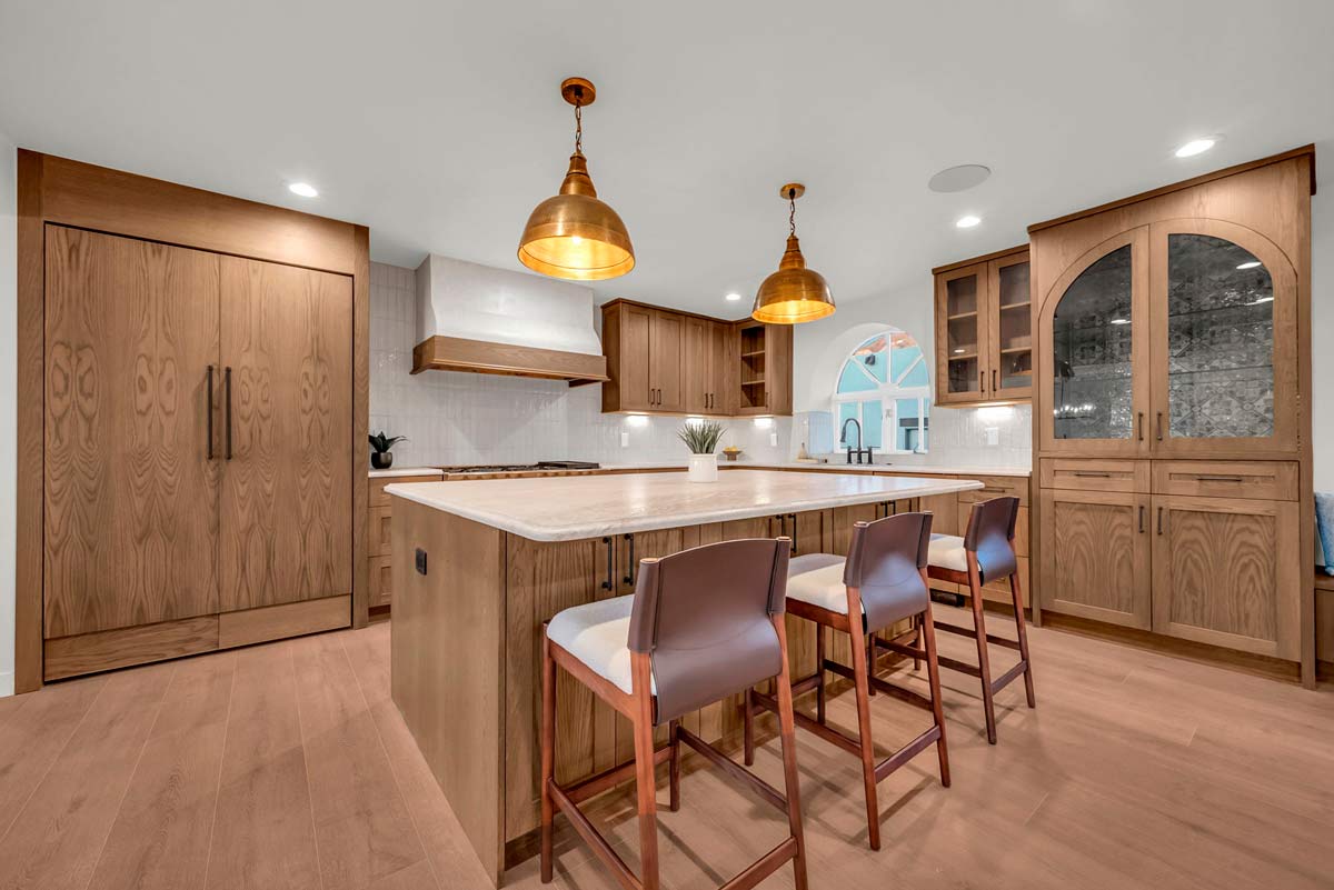 A modern kitchen with wooden cabinets and floors, featuring a large island with four bar stools. Two copper pendant lights hang above the island. A built-in fridge and glass-front cabinet are visible in the background.
