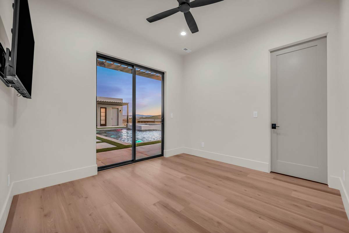 Minimalist room featuring wooden flooring, a wall-mounted TV, and a ceiling fan. A sliding glass door opens to a view of a pool and pool house with a scenic sunset in the background. A closed door is on the right wall.