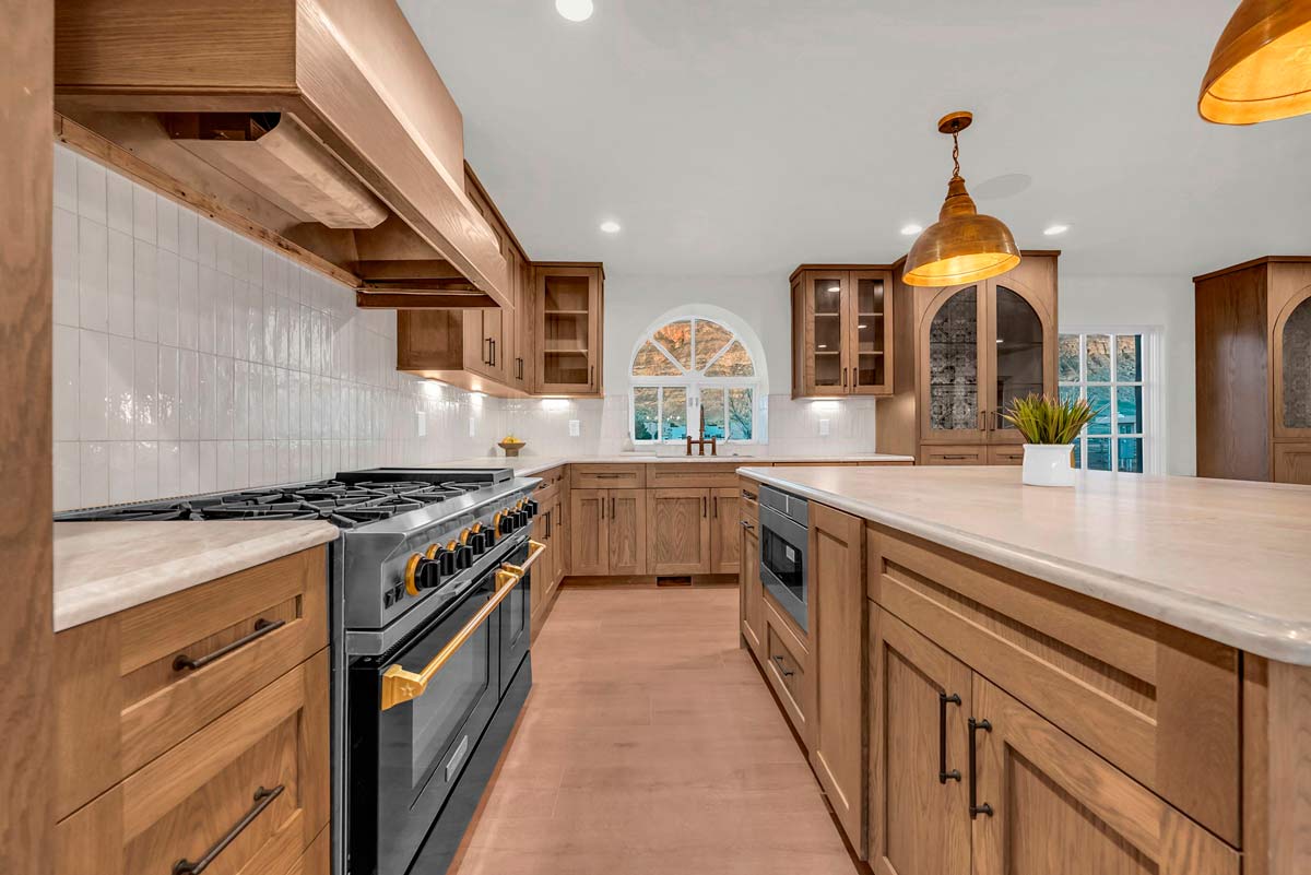 A modern kitchen with wooden cabinets and a large island. The space features a black stove, two pendant lights, and a small potted plant on the island. A white tiled backsplash and an arched window add elegance to the room.