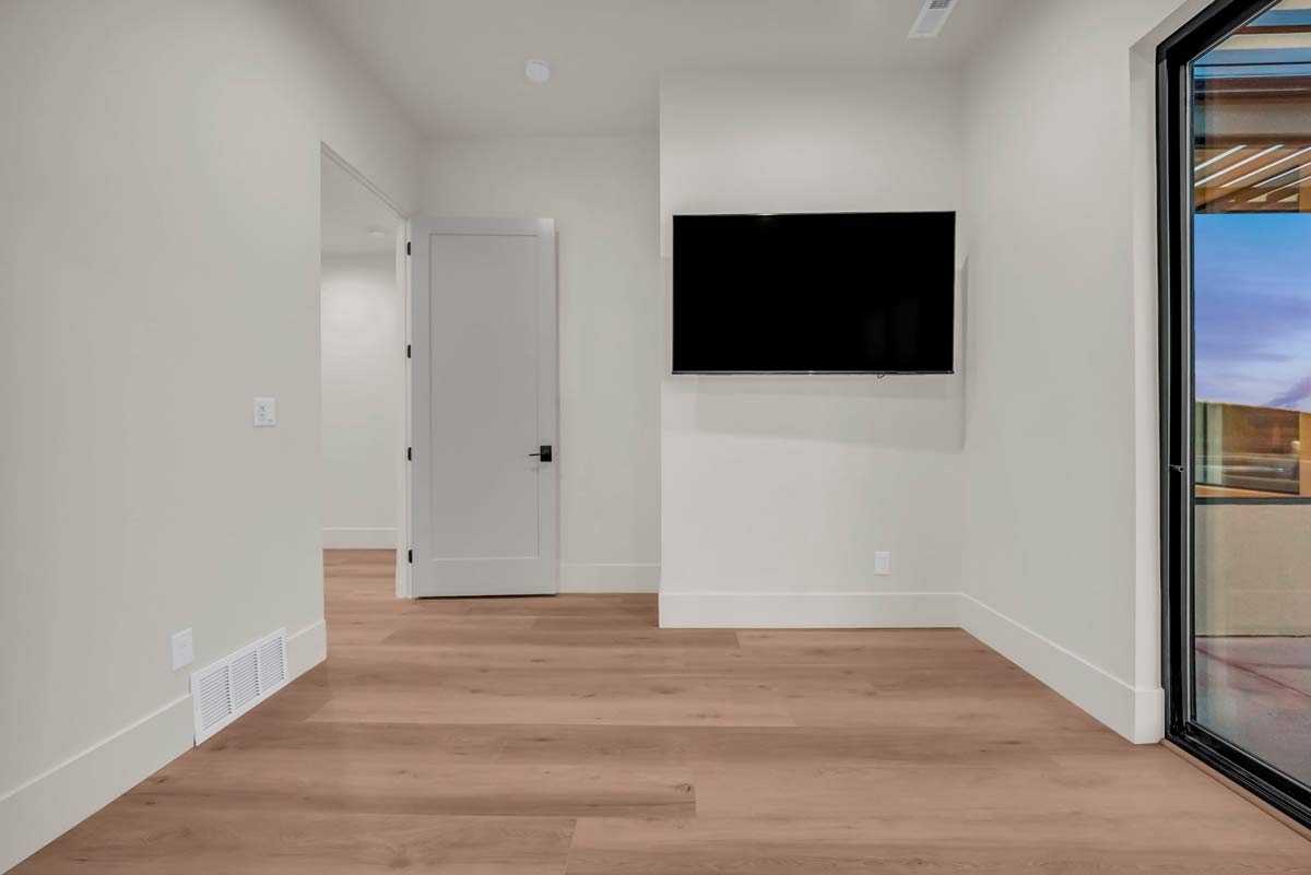 Minimalist room with light wood flooring and white walls. A flat-screen TV is mounted on the wall. The room has an open door to the side and a large sliding glass door leading to an outdoor area.