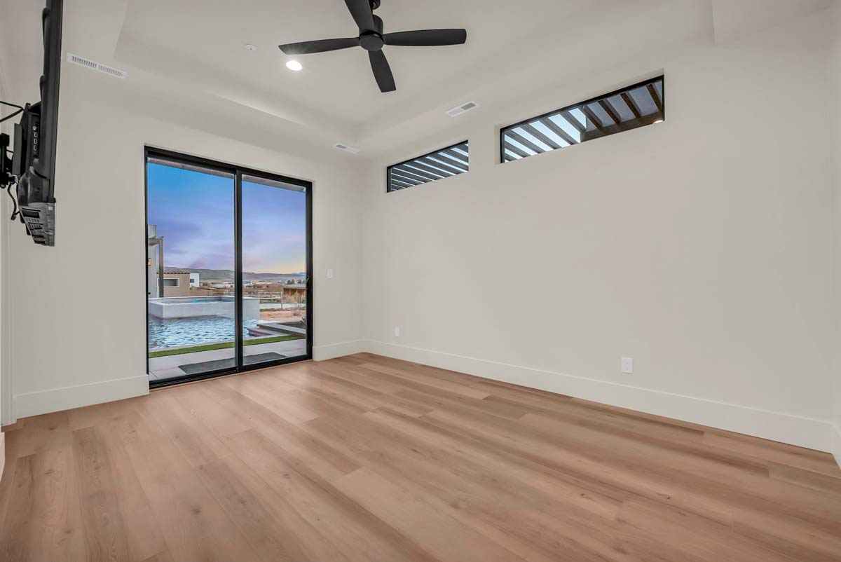 Bright, empty room with light wood flooring, beige walls, and a ceiling fan. Large sliding glass doors open to a pool view, and small windows near the ceiling let in additional light.