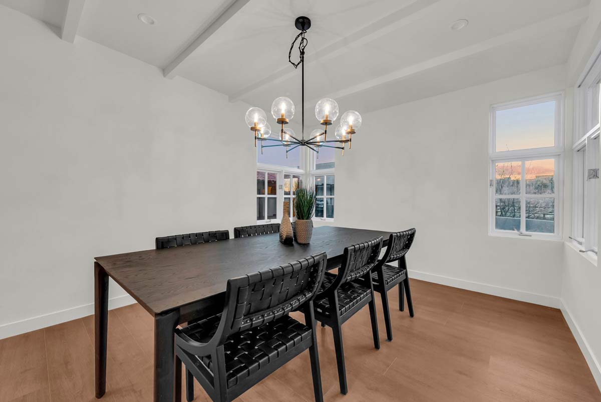 Dining room with a dark wooden table and six woven black chairs. A modern chandelier with seven glass globes hangs above. Large windows allow natural light, illuminating the light-colored walls and wooden floor.