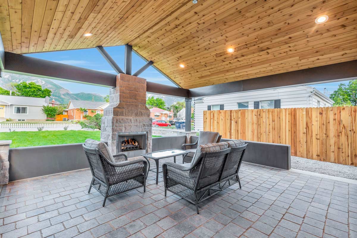 Covered patio with a wooden ceiling and recessed lights, featuring a stone fireplace. Four wicker chairs surround a small table on a tiled floor. In the background, a wooden fence and suburban houses are visible.