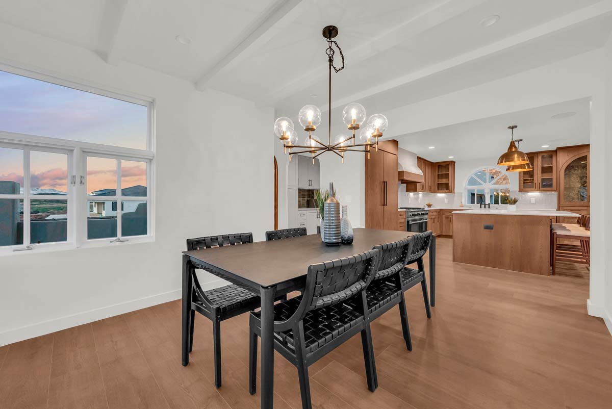 A modern dining area with a black table and six woven chairs. A stylish chandelier hangs above. Beyond the dining room is a spacious kitchen with wooden cabinets and a central island. Large windows show a scenic view outside.