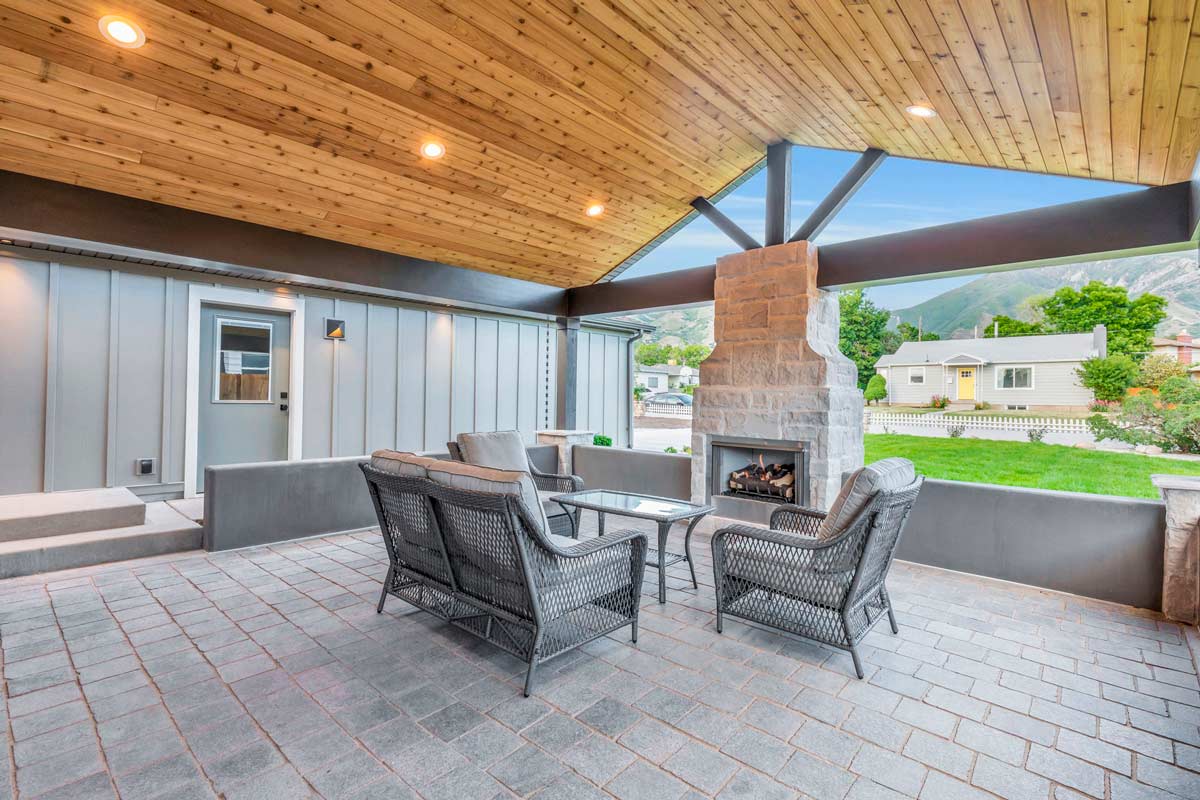 Covered outdoor patio with a wooden ceiling and recessed lighting. Features wicker chairs and a stone fireplace. Overlooks a grassy yard with trees and houses in the background, framed by distant mountains.