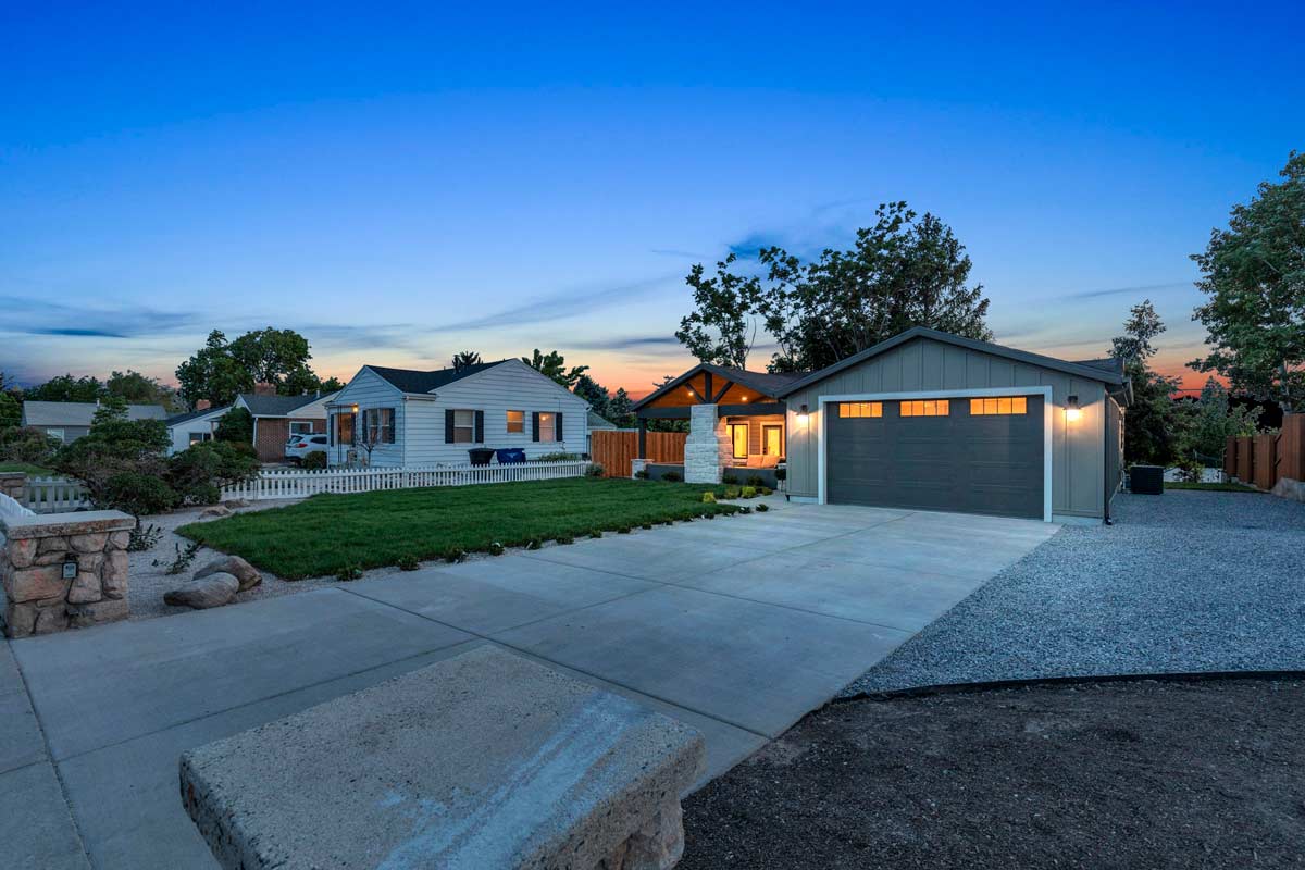 A modern house with a large driveway and attached garage is shown at dusk. The home has illuminated windows and a front yard with green grass. Another house is visible nearby, and trees surround the area under a twilight sky.