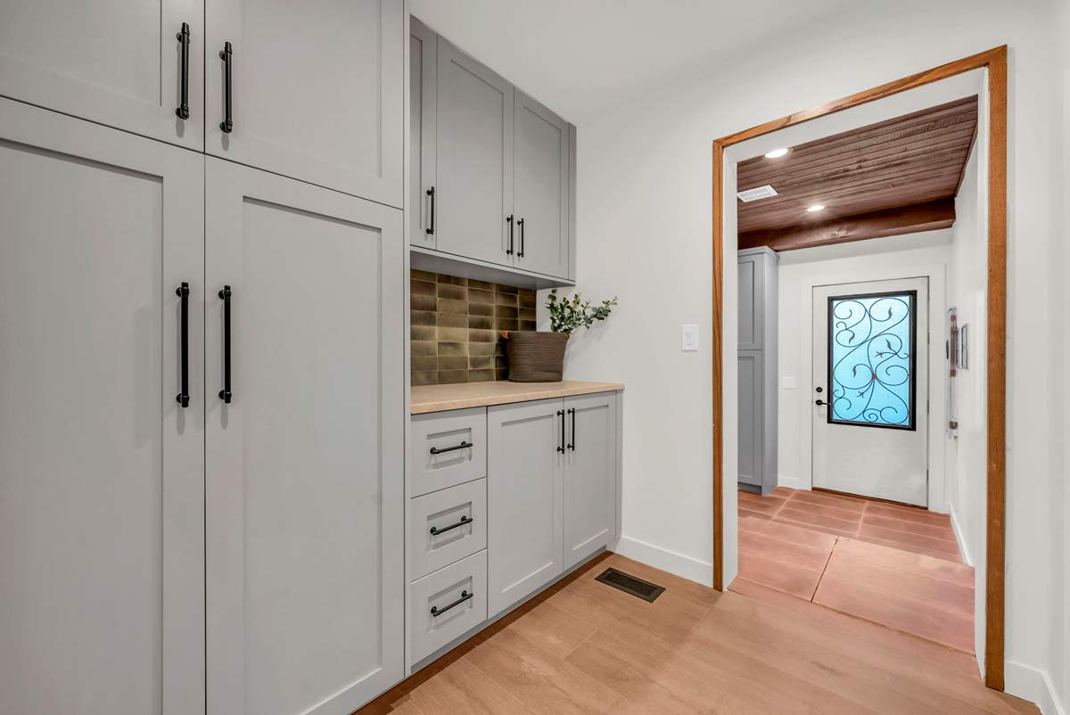 A hallway with light gray cabinets on the left, featuring black handles. Theres a wooden countertop with a pot of greenery. The walls are white, and the floor is wooden, leading to a door with an ornate glass design.