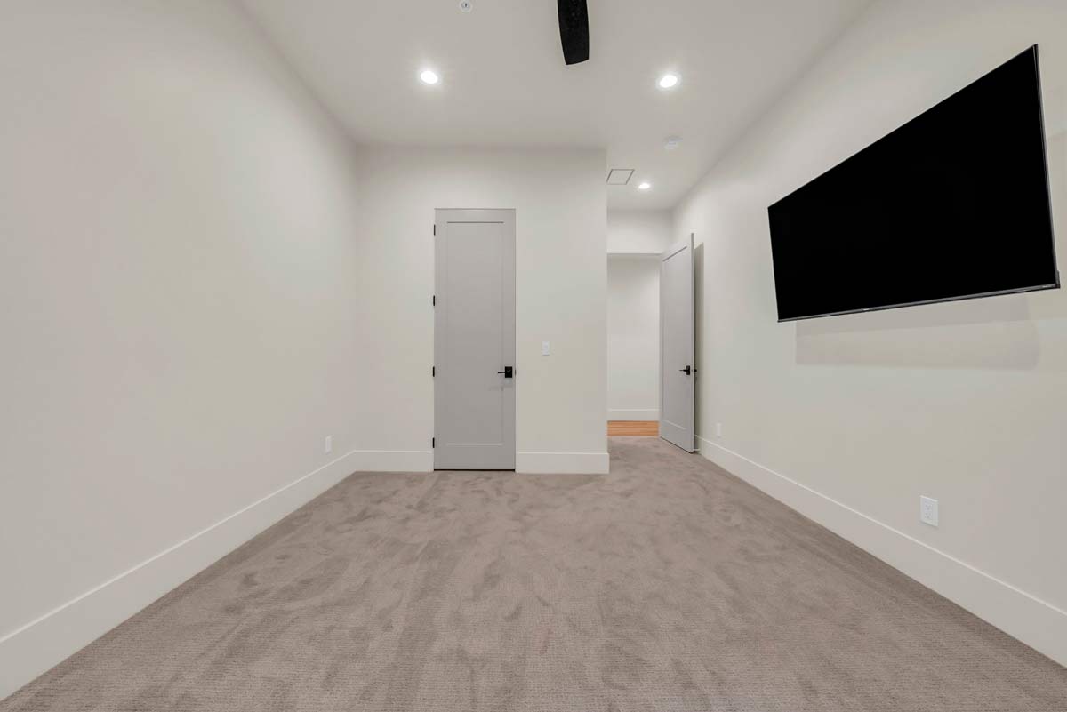 A sparse room with beige walls and carpet, featuring a closed gray door on one wall and a mounted flat-screen TV on another. A slightly open door leads to a lit hallway with wooden flooring. Ceiling lights illuminate the space.