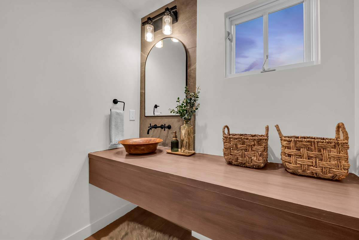Minimalist bathroom featuring a floating wooden vanity with two woven baskets and a wooden bowl. An arched mirror with two pendant lights hangs above, adjacent to a small window showing a sunset sky. A towel is on a nearby holder.