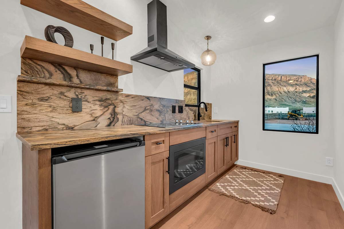 Modern kitchen with wood cabinetry, a stone countertop and backsplash, and a small fridge. A stovetop and oven are built-in, with open wooden shelves above. A pendant light hangs from the ceiling, and a window offers a scenic mountain view.