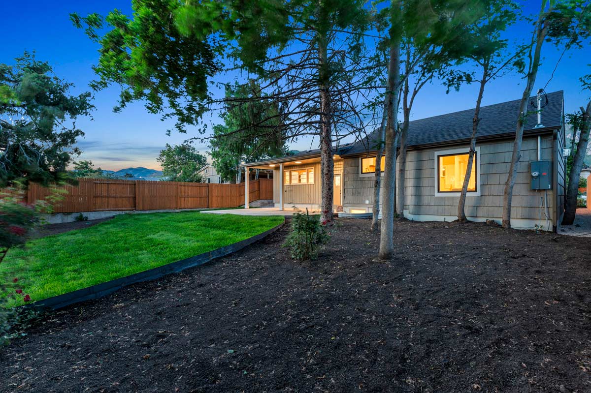 A small house with beige siding, lit from the inside, stands surrounded by a freshly landscaped yard. Theres a lush green lawn, newly planted trees, and a wooden privacy fence, all under a clear blue twilight sky.