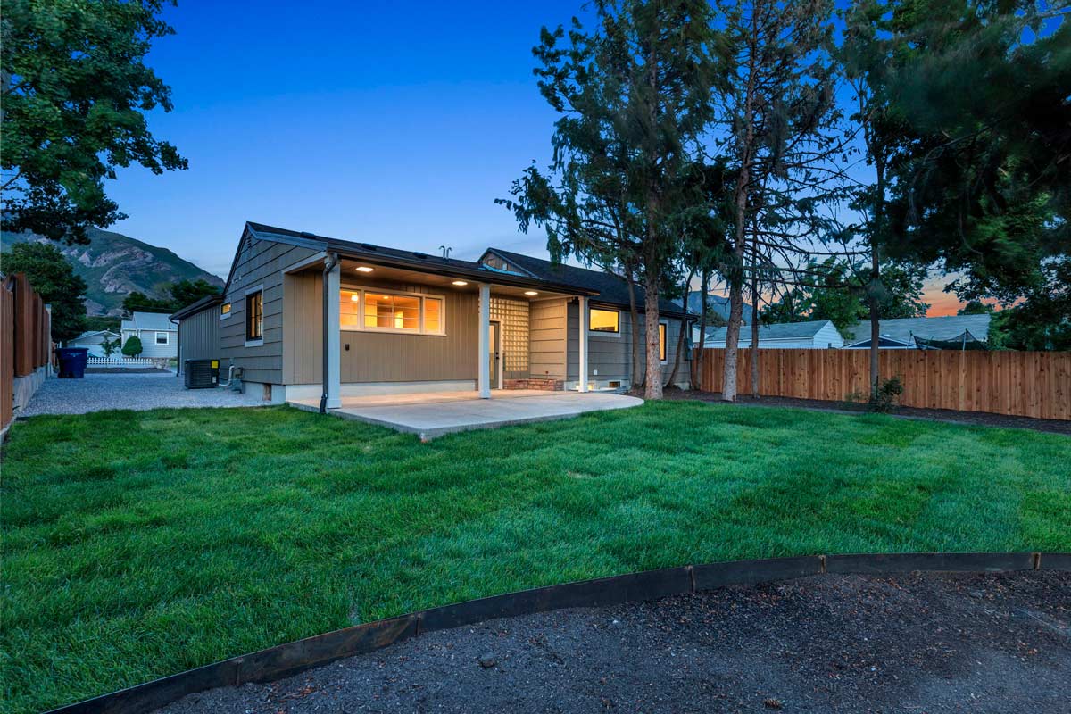A modern, single-story house with a small covered patio is surrounded by a grassy backyard. Tall trees line the fenced area. The sky is clear at dusk, creating a peaceful atmosphere.