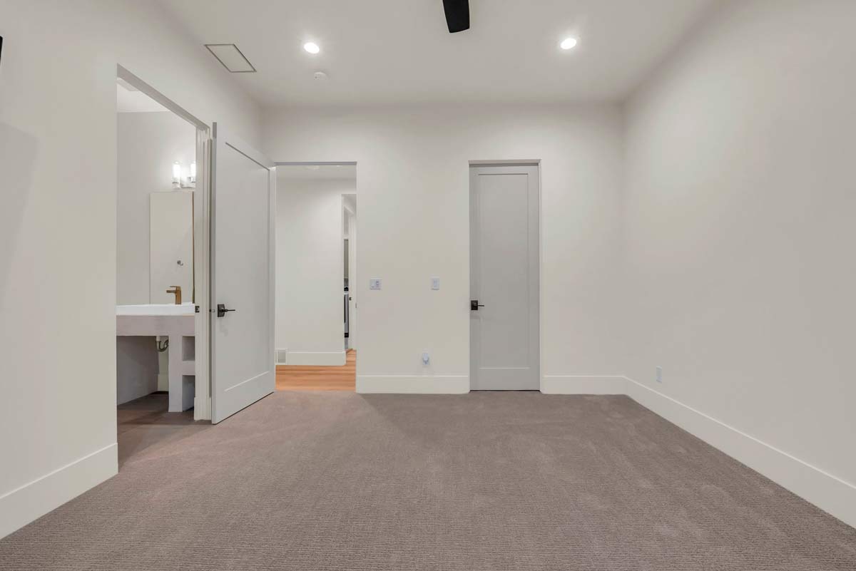 Empty room with beige carpet and white walls. Two doors are closed, and one door is partially open, revealing a bathroom with a sink and mirror. Recessed lighting is installed in the ceiling.