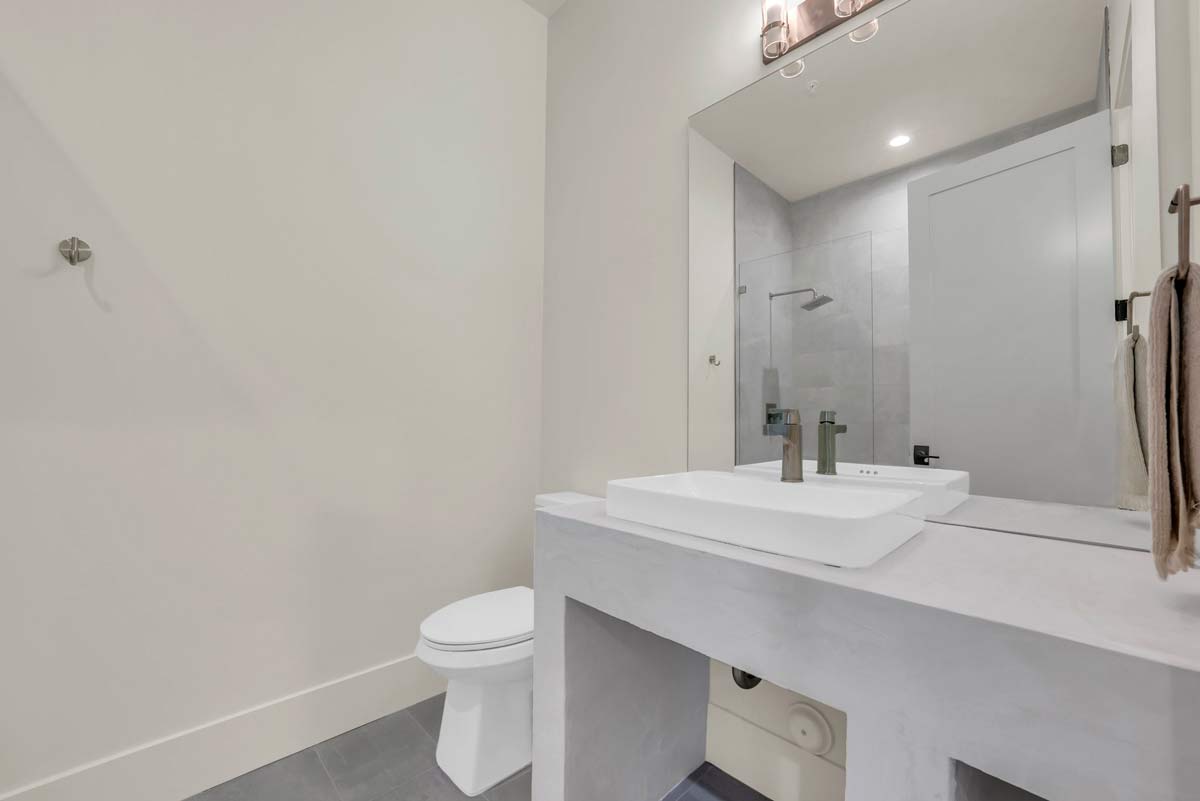 Modern bathroom with light gray walls and flooring, featuring a sleek rectangular white sink on a minimalist vanity. A large mirror spans the wall above the sink. A towel hangs on the right, and a toilet is positioned on the left.