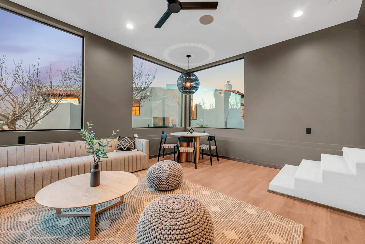 Modern living room with large windows, a corner dining area, and light wood flooring. Room features a gray sectional sofa, round poufs, a wooden coffee table, and a black pendant light. White stairs lead up to the right.