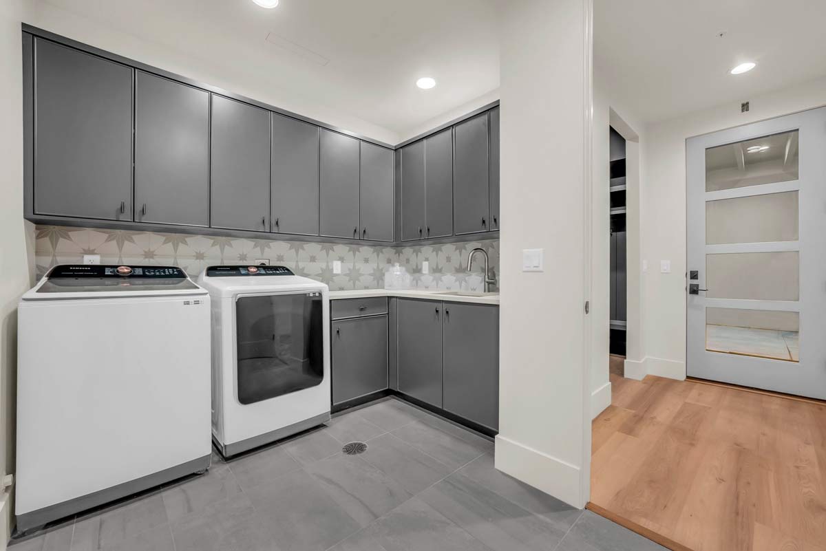 A modern laundry room with gray cabinets and a countertop above a white washing machine and dryer. The room has tiled flooring, a patterned backsplash, and a glass-paneled door leading to a hallway with wooden flooring.